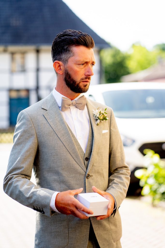 Mann in grauem Anzug mit Fliege hält eine kleine Schachtel, Blick nachdenklich.