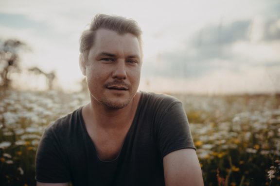 Junger Mann mit dunklem Shirt in einer Blumenwiese, sanftes Licht im Hintergrund.