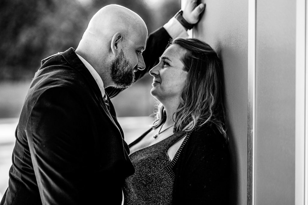 Paar in formeller Kleidung schaut sich verliebt an, mit einer Wand im Hintergrund.