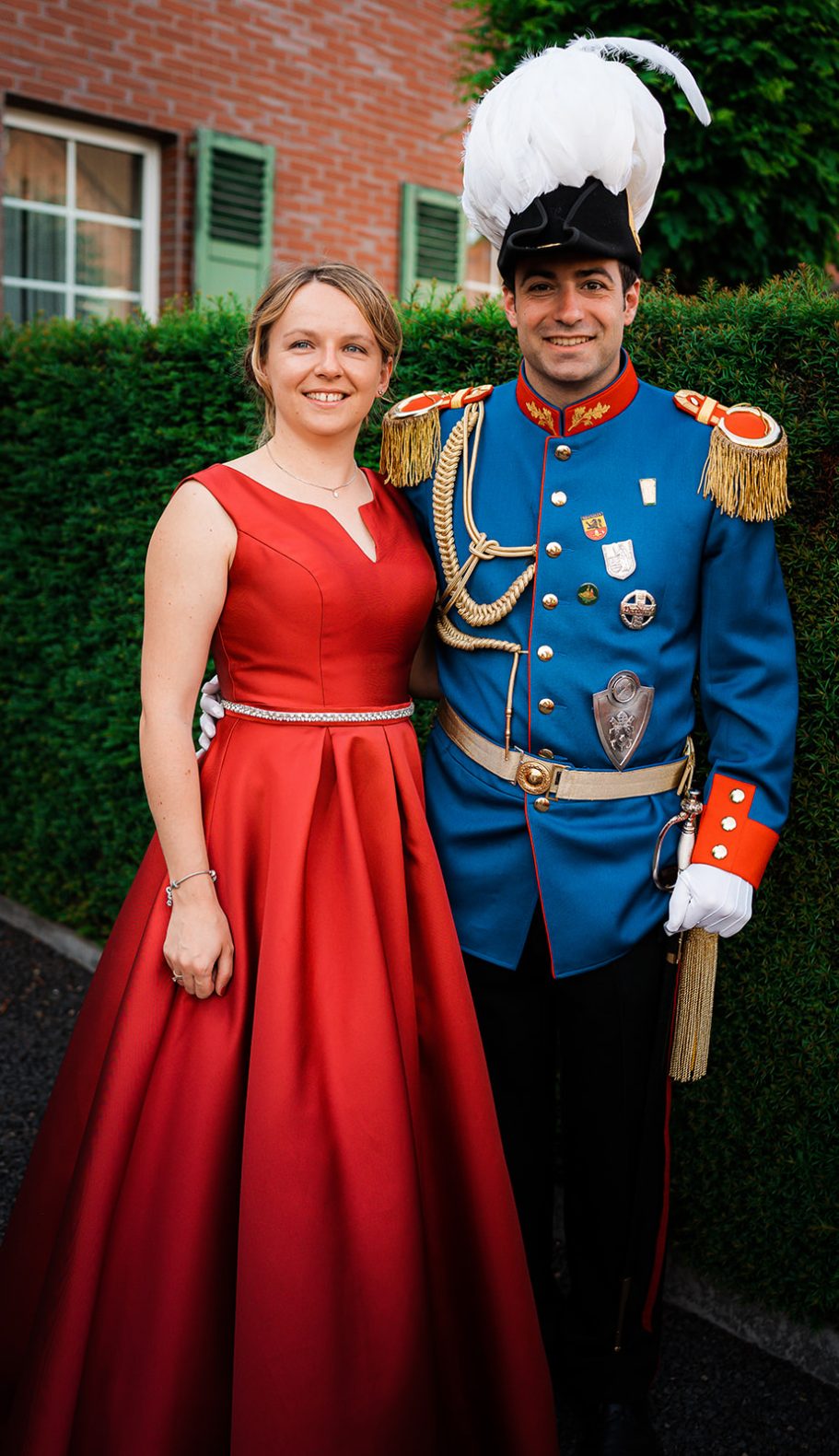 Ein Paar in festlicher Kleidung, die Frau in einem roten Kleid, der Mann in uniformähnlicher Kleidung.
