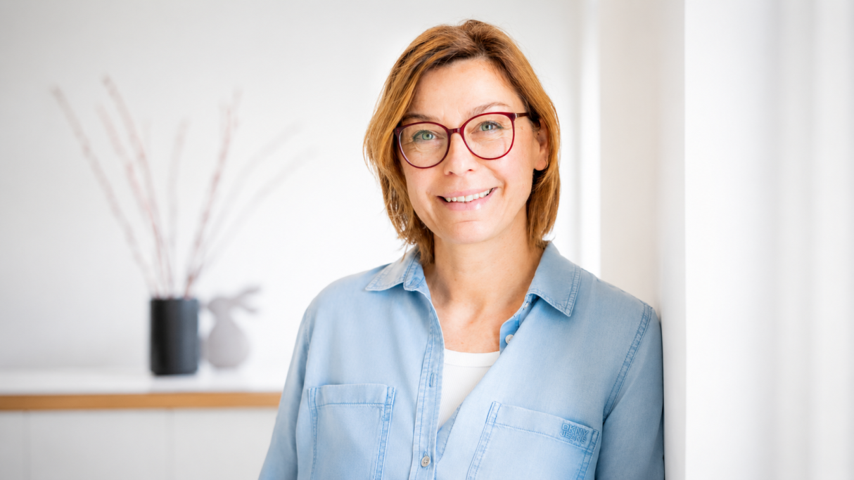 Frau mit Brille und blauer Bluse, lächelt vor heller Wand und Vasen im Hintergrund.