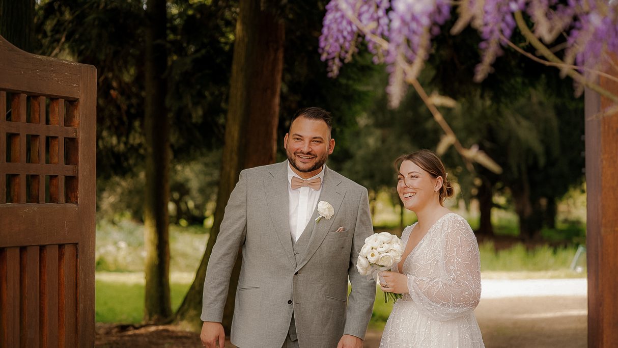Brautpaar steht lächelnd vor einer Blütenpracht in einem Garten.