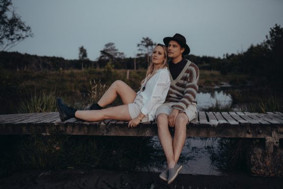 Zwei Personen sitzen auf einer Holzbrücke in einer naturbelassenen Umgebung.