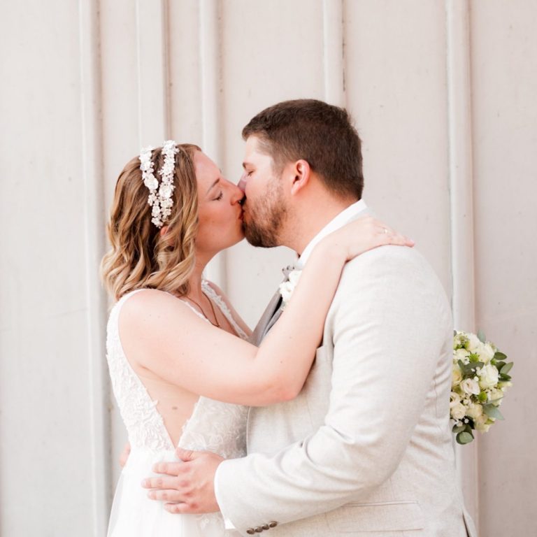Ein Brautpaar küsst sich vor einer neutralen Wand, umgeben von Blumen.