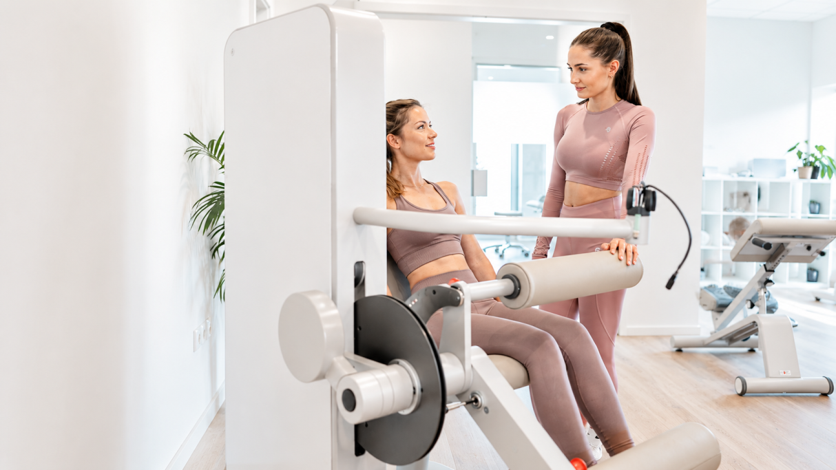 Zwei Frauen im Fitnessstudio, eine trainiert an einem Trainingsgerät, die andere gibt Anleitung.