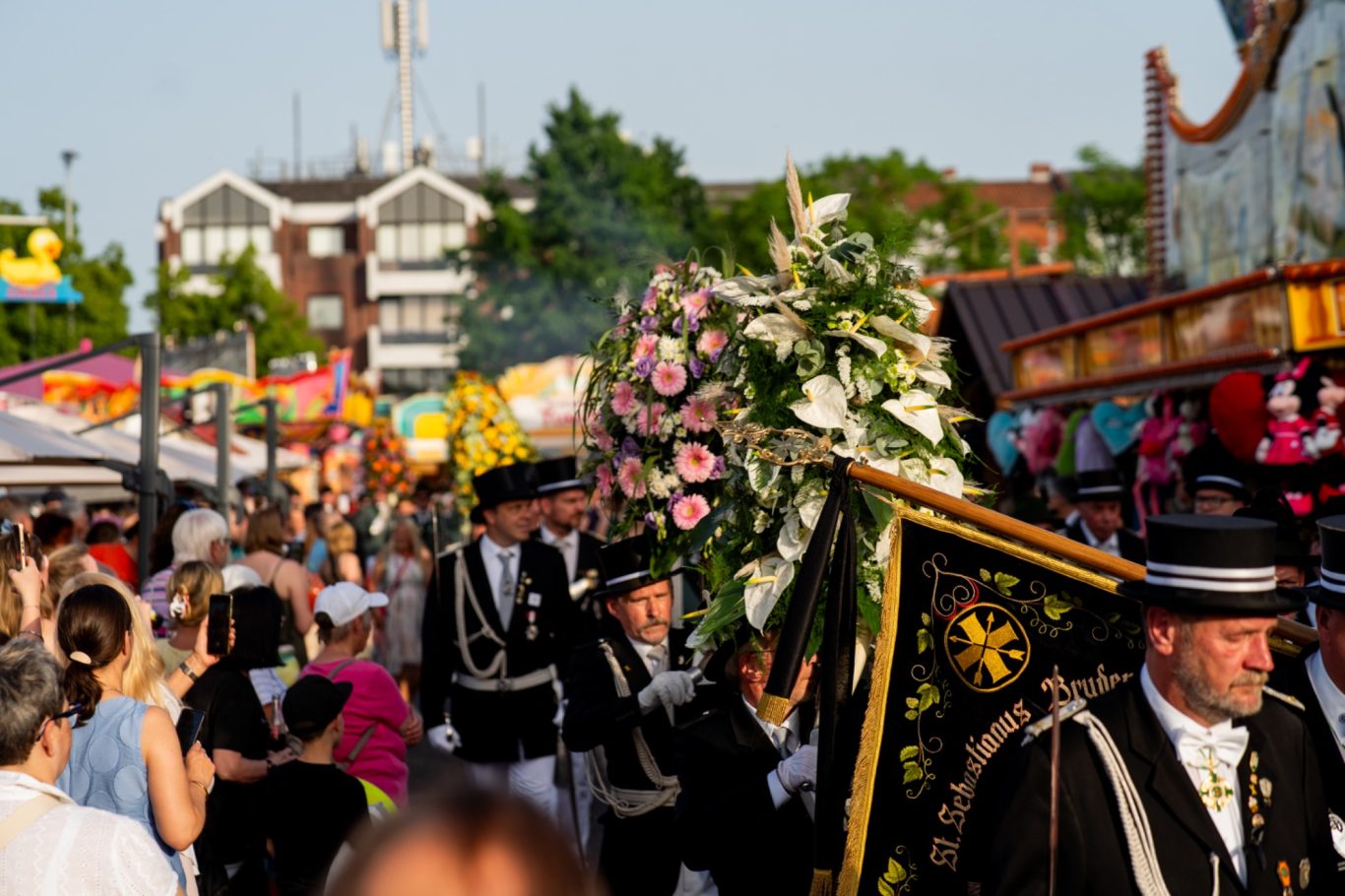 Menschenmenge auf einem Festival, mit Darstellern in festlicher Kleidung und Blumenarrangements.