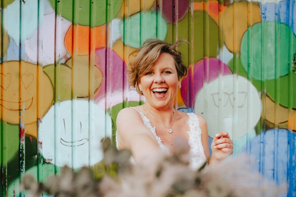 Lachende Frau vor buntem, lächelndem Wandgemälde mit Blumen im Vordergrund.