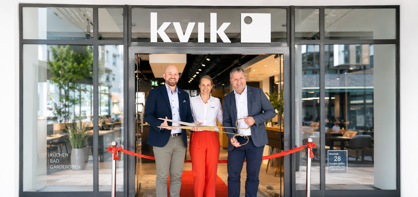 Drei Personen vor einem Kvik-Shop, halten Auszeichnungen, mit rotem Band im Hintergrund.