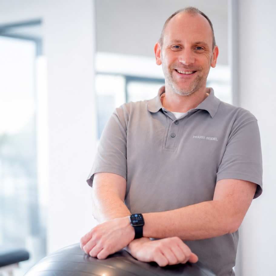 Mann in grauem Poloshirt lächelt und stützt sich auf einen Ball.