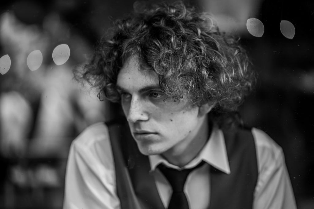 Junger Mann mit lockigem Haar, nachdenklichem Ausdruck und eleganter Kleidung.