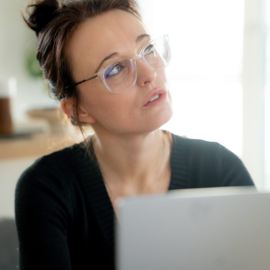 Eine nachdenkliche Frau mit Brille sitzt vor einem Laptop.