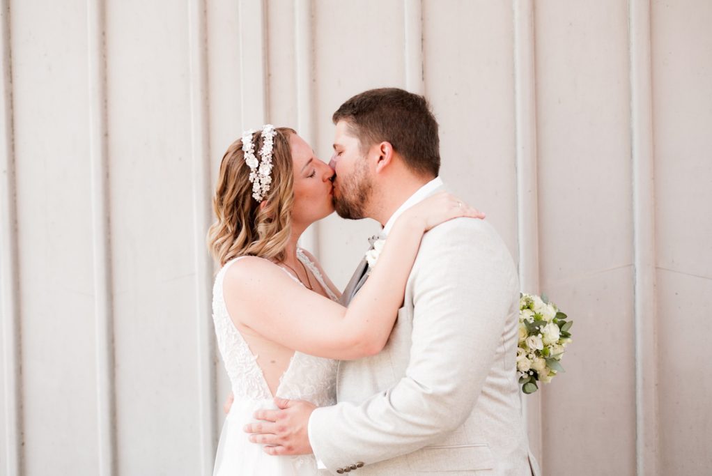 Ein Brautpaar küsst sich innig vor einer hellen Wand.