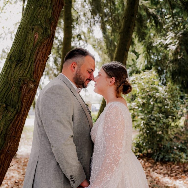 Brautpaar steht sich gegenüber, ihre Stirnen berühren sich, umgeben von Bäumen.