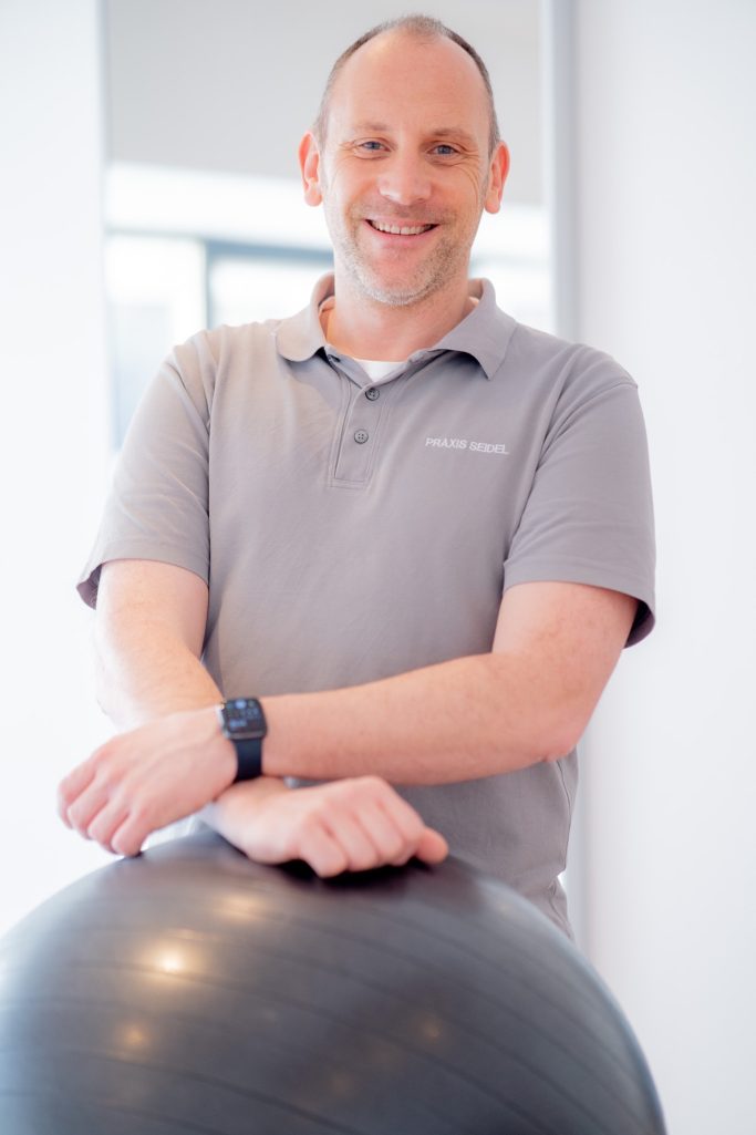 Mann mit sportlichem Oberteil steht neben einem Gymnastikball und lächelt freundlich.