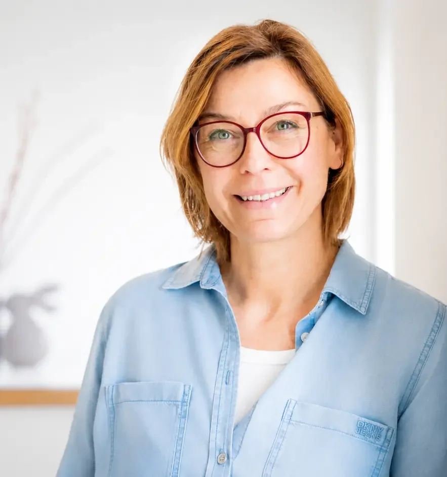 Frau mit kurzen braunen Haaren und Brille, lächelt freundlich in heller Umgebung.